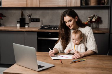 Mom and toddler daughter draw pictures with colored pencils from laptop. Developing drawing lesson for child toddler at home. Maternity leave