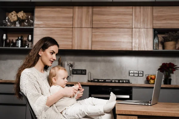Mom and child daughter are watching cartoons on laptop at home in the kitchen. Motherhood. Family of mother and daughter toddler spending time together and watching videos online