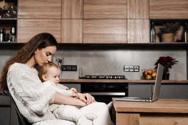 Family of mother and daughter toddler spending time together and watching videos online. Motherhood. Mom and child daughter are watching cartoons on laptop at home in the kitchen
