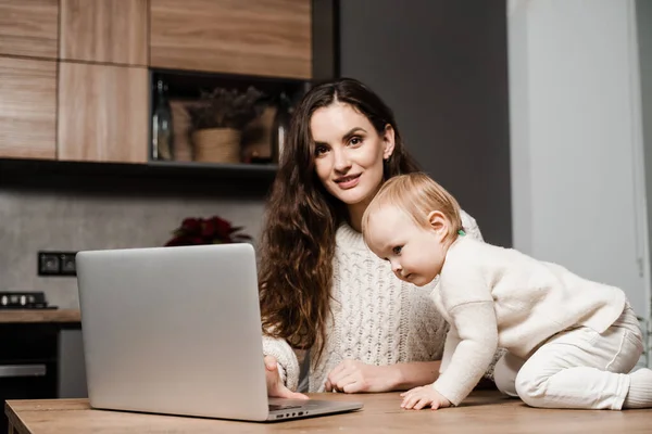 Mom and child daughter are watching cartoons on laptop at home in the kitchen. Motherhood. Family of mother and daughter toddler spending time together and watching videos online