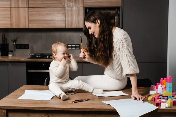 Mom and daughter toddler fool around and play together in the kitchen at home. Happy family of mom and daughter laugh and play with colored pencils while drawing. Maternity leave