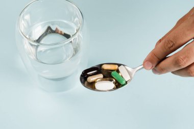 Pills on spoon of dietary biologically active supplements and glass of water for daily tablets intake on blue background. Set of omega and zinc pills, antioxidants from aging, lecithin and adaptogen