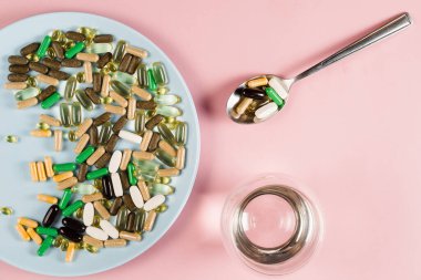 Many dietary supplements tablets, glass of water and spoon for daily pills intake on pink background. Set of zinc pills, antioxidants from aging, lecithin and adaptogen