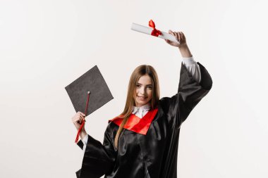 Happy student in black graduation gown and cap is smiling on white background. Graduate girl is graduating high school and celebrating academic achievement. Masters degree diploma in hands