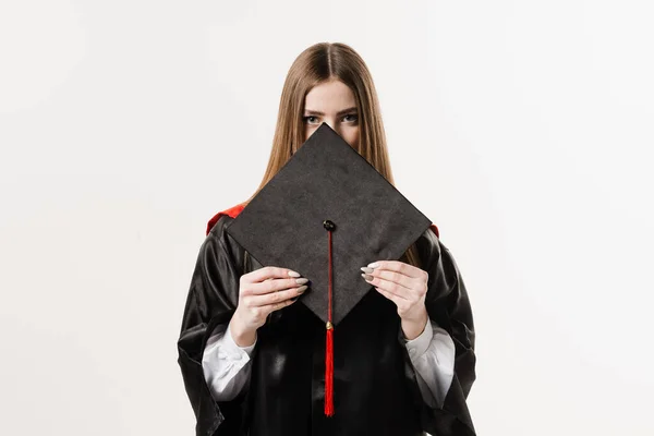 Graduate girl master degree in black graduation gown is holding cap in hands on white background. Attractive young woman graduated from college