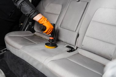 Applying detergent on textile seat in car interior for dry cleaning. Smearing detergent on car textile seats using drill with brush for dry cleaning