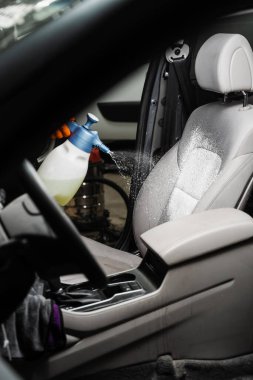 Spraying detergent on textile seats in car interior for dry cleaning. Professional cleaner in gloves is pouring detergent on seat in car for removing stains and dirt