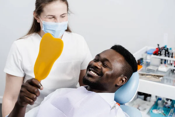 African man looking on teeth in the mirror at the dentist. Happy african american patient of dentistry. Teeth treatment. Handsome man sitting in dental office and smiling