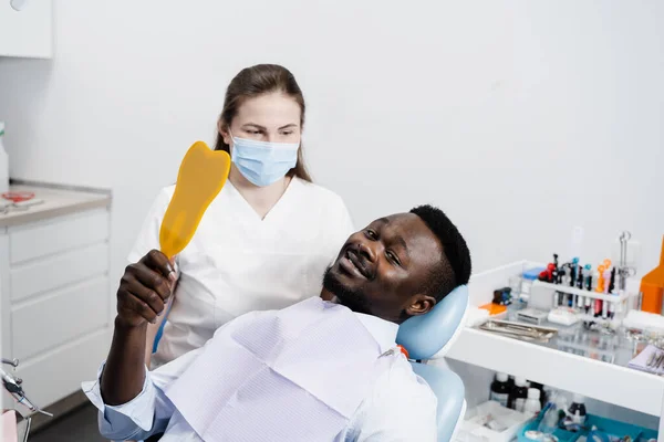 African man looking on his teeth in the mirror at the dentist. Happy african american patient of dentistry. Teeth treatment. Handsome man sitting in dental office and smiling