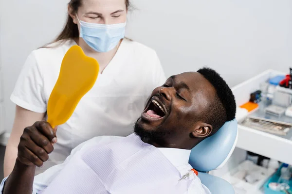 African man looking on his teeth in the mirror at the dentist. Happy african american patient of dentistry. Teeth treatment. Handsome man sitting in dental office and smiling