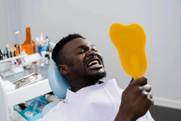 Consultation with dentist in dental clinic. African american patient is visiting dentist. African man patient is looking in the mirror at his teeth after removing caries and filling teeth
