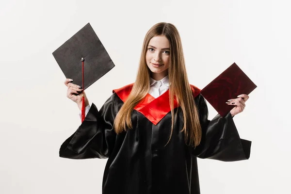 Graduate girl master degree in black graduation gown is holding diploma and cap in hands on white background. Attractive young woman graduated from college