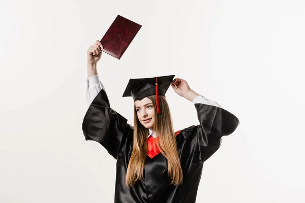 Happy girl student in black graduation gown and cap raises masters degree diploma above head on white background. Graduate girl is graduating college and celebrating academic achievement