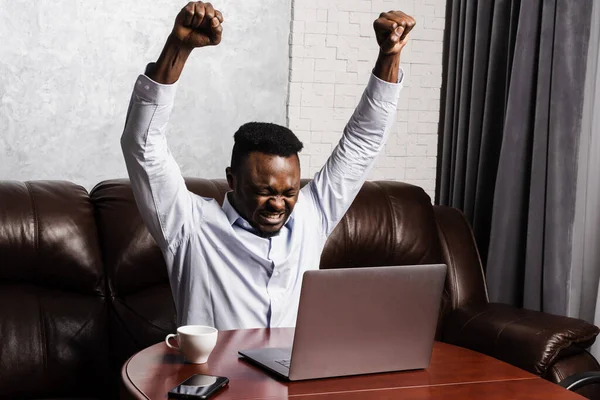 Happy cheerful african american man with laptop raise hands up and smile and looking at amount of income earnings and rejoices. Happy african man rejoices in winning and sitting in front of laptop