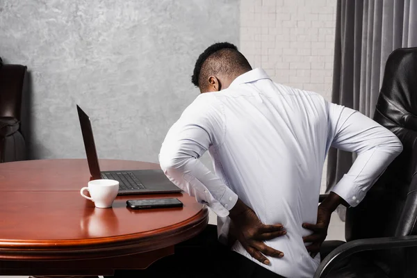 African man feels back pain after working for long hours with laptop online in office. Cervical spine osteochondrosis is radicular syndromes of african american man