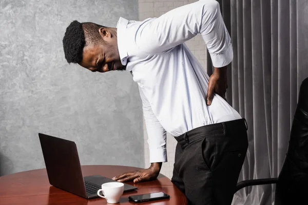 African man feels back pain after working for long hours with laptop online in office. Cervical spine osteochondrosis is radicular syndromes of african american man