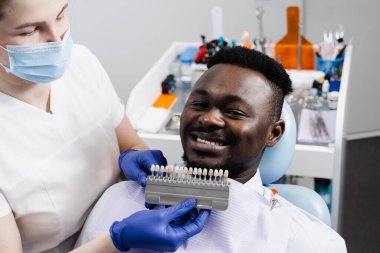 Afrikalı adamın dişlerinin beyazlaması. Diş hekimi, diş tonu rehberiyle çalışıyor. Diş hekimliği. Doktor diş sağlığı kliniğinde diş rengine uyan örnekleri kontrol ediyor.