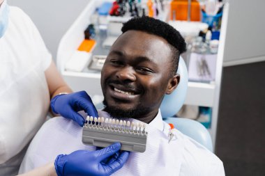 Afrikalı adamın dişlerinin beyazlaması. Diş hekimi, diş tonu rehberiyle çalışıyor. Diş hekimliği. Doktor diş sağlığı kliniğinde diş rengine uyan örnekleri kontrol ediyor.