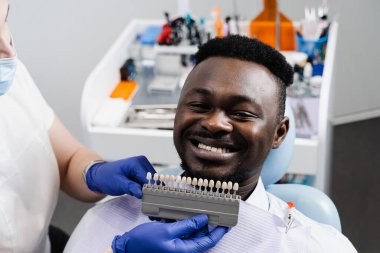 Diş hekiminde Afrikalı hastalara diş beyazlatma için diş rengi tonları gösteren bir diş hekimi. Afrikalı, kaplamalara bakıyor ya da diş implantları yerleştiriyor. Doktorun ellerine eşleşen örnekler. Dişçilik