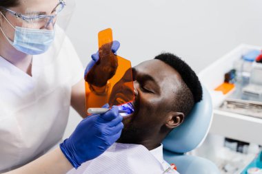 Afrikalı diş hekimi için fotopolimer diş dolgusunun UV aydınlatması. Koruyucu gözlüklü diş hekimi Afrikalı hastaları tedavi edip çıkarıyor.