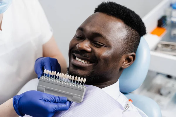 Teeth whitening of african man. Dentist working with teeth color shades guide. Dentistry. Doctor checking teeth color matching samples in dental clinic