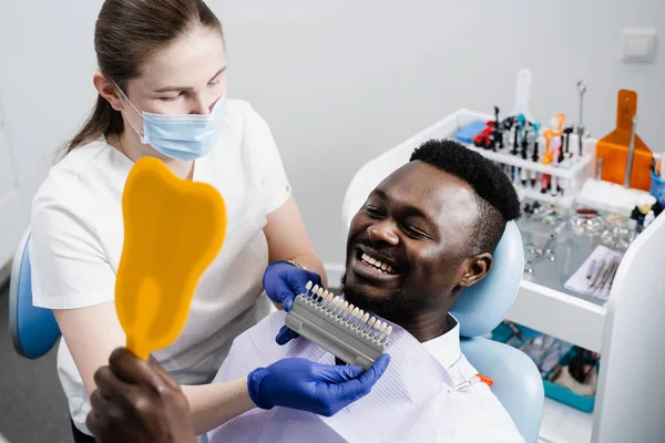 African is looking at veneers or implants teeth color matching samples in doctor hands. Dentistry. Dentist showing teeth color shades guide for tooth whitening for african patient in dental clinic
