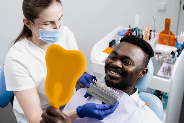 African is looking at veneers or implants teeth color matching samples in doctor hands. Dentistry. Dentist showing teeth color shades guide for tooth whitening for african patient in dental clinic