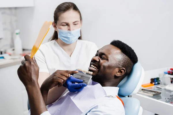 Dentistry. Doctor checking teeth color matching samples in dental clinic. Teeth whitening of african man. Dentist working with teeth color shades guide