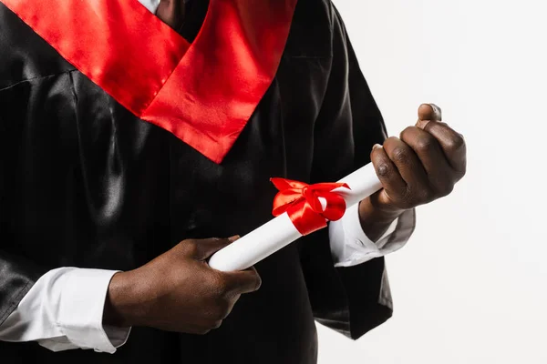 Masters degree diploma with red ribbon in hands of graduate african man in black graduation gown on white background. Graduate african is graduating high school and celebrating academic achievement