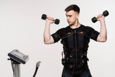 EMS giysisi içinde dambılları olan kaslı erkek sporcu beyaz arka plandaki kasları uyarmak için elektriksel dürtüler kullanıyor. Elektrik kas stimülasyonu giysisinde spor eğitimi