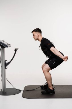 Elektrik kas stimülasyonu giysisinde spor eğitimi. EMS takım elbiseli kaslı erkek sporcu, beyaz arka plandaki kasları uyarmak için elektriksel dürtüler kullanan spor eğitimi yapıyor.