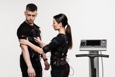 Kadın ve erkek, beyaz arka plandaki kasları uyarmak için elektriksel impulsları kullanan EMS giysisinin kontaktörlerini birbirine bağlar. Elektrik kas stimülasyonu için spor grubu eğitimi