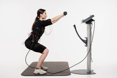 Formda kız, beyaz arka plandaki kasları uyarmak için elektirikli impulsları kullanan EMS giysisiyle halterli fiziksel egzersizler yapıyor. Elektrik kas stimülasyonu giysisinde spor eğitimi