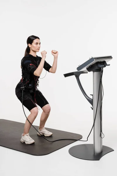 Stock image Fit girl does squats physical exercises in EMS suit that uses electrical impulses to stimulate muscles on white background. Sport training in electrical muscle stimulation suit