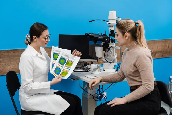 Diagnosis of the cornea and eyes in ophthalmology clinic. Ophthalmologist is showing keratotopography scan to girl patient near slit lamp