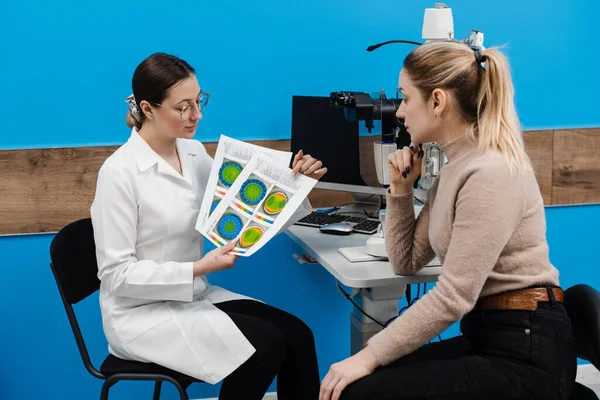 Diagnosis of the cornea and eyes in ophthalmology clinic. Ophthalmologist is showing keratotopography scan to girl patient near slit lamp