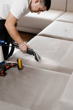 Temizlikçi, elektrikli süpürge makinesiyle mobilyaları temizlemek için kanepeyi temizliyor. Temizlikçi kuru temizleme makinesiyle döşenmiş koltuktan toprak çıkarıyor.