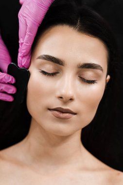 Taş kazıyıcı kullanan bir kız yüzü için guaj masajı. Güzellik uzmanı Gua Sha masajı yapar kan dolaşımını ve yüz rengini iyileştirmek için.