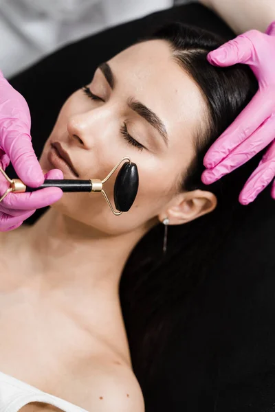 Cosmetologist using stone roller for face massage for girl patient. Facial massage with cosmetics roller in cosmetology clinic. Process of face massage with black stone roller close-up