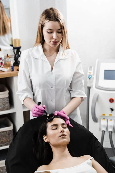 Process of face massage with black stone roller. Cosmetologist using stone roller for face massage for girl patient. Facial massage with cosmetics roller in cosmetology clinic