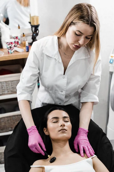 Process of face massage with black stone roller. Cosmetologist using stone roller for face massage for girl patient. Facial massage with cosmetics roller in cosmetology clinic