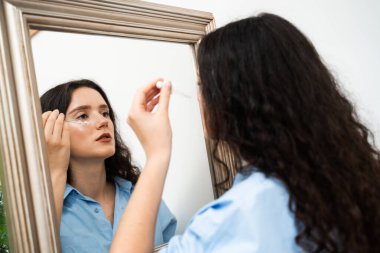 Çekici kız evdeki aynada yüz derisine gerekli yağı döküyor. Genç bir kadın yüzüne nemlendirici serum sürüyor.