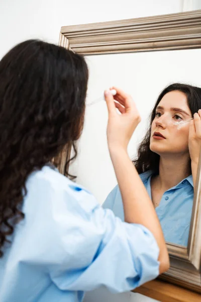 stock image Young woman is applying moisturizing serum on skin face is looking in the mirror. Girl with hyaluronic acid or serum pipette in hands close-up