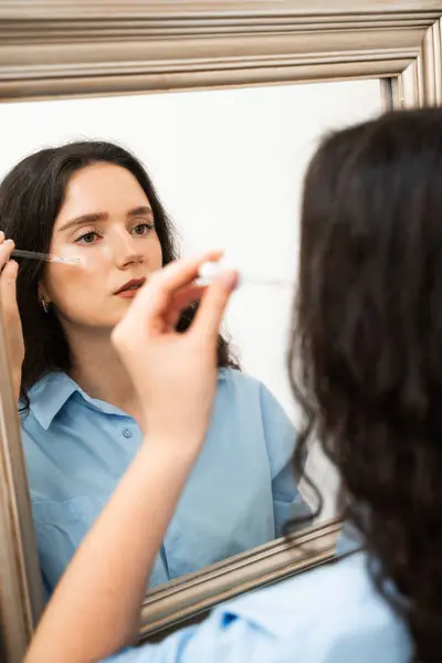 stock image Attractive girl drops essential oil on facial skin in the mirror at home. Young woman is applying moisturizing serum on her face