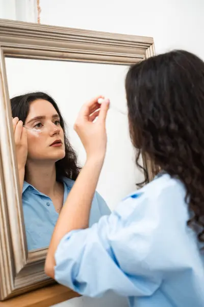 stock image Young woman is applying moisturizing serum on skin face is looking in the mirror. Girl with hyaluronic acid or serum pipette in hands close-up