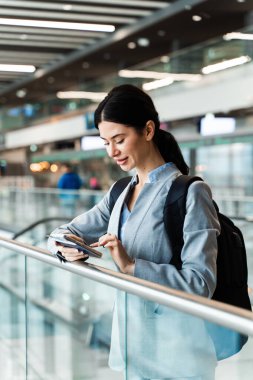 Havaalanı terminalinde yürüyen merdivenin yanında belgelerle bekleyen gezgin 2024 yaz tatilinin başlamasını bekliyor. Havaalanı lobisindeki akıllı telefondan bakan kadın..