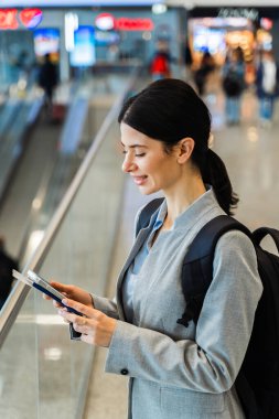Havaalanı salonunda akıllı telefon, belge ve sırt çantalı bir turist panoramik pencereden bakıyor. Uçuşun 2024 'te başlamasını bekleyen gülümseyen beyaz kadın.