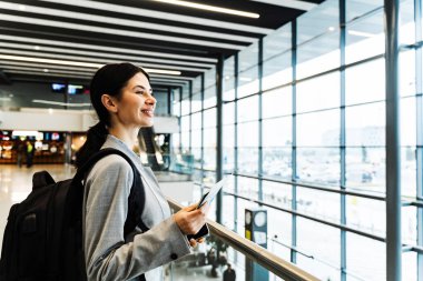 Havaalanı salonunda akıllı telefon, belge ve sırt çantalı bir turist panoramik pencereden bakıyor. Uçuşun 2024 'te başlamasını bekleyen gülümseyen beyaz kadın.