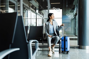 Mavi bavulun sapı kadının elinde ve havaalanı terminaline bakıyor. İş kadını havaalanında oturuyor ve seyahatine başlamayı bekliyor.