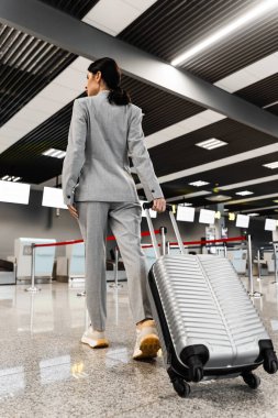 Bir kadın havaalanı terminalindeki güvenlik kontrolünden bavulunu geçiriyor. Havaalanı lobisinde gri bavullu bir turist.